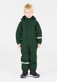 Groene waterdichte overall met een rits aan de voorkant, elastische manchetten en reflecterende accenten. Gecombineerd met zwarte waterdichte laarzen.