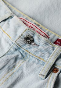 Jasnobrązowy pas do niebieskich dżinsów z widocznym metalowym guzikiem oznaczonym napisem „Superdry Denim Goods” i czerwoną metką marki wewnątrz.