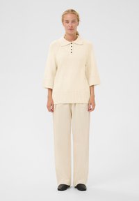 Pull oversized beige clair avec col polo, trois boutons et poignets côtelés, associé à un pantalon large beige pâle et des chaussures noires.