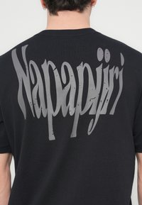 Črna bombažna majica s kratkimi rokavi in okroglim izrezom, na hrbtni strani pa ima velik, raztrgan siv napis »Napapijri«.