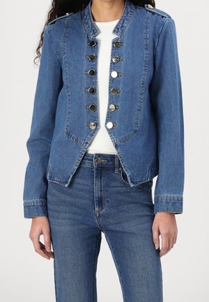 Vrouw draagt een blauw spijkerjack met zilveren knopen over een wit topje en blauwe spijkerbroeken tegen een effen achtergrond.