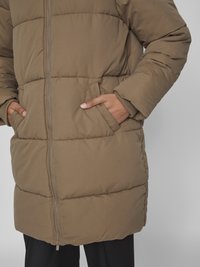 Brun vadderad jacka med dragkedja framtill, sidofickor, ribbade ärmslut och kviltat mönster. Tyget verkar mjukt och isolerat för värme.