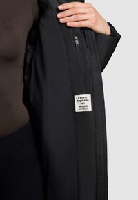 Person, der eine schwarze Jacke trägt, mit einem inneren Reißverschlussfach und einem Stoffetikett mit der Aufschrift "Eine warme Umarmung aus Stoff."