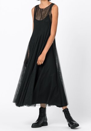 Personne portant une robe midi noire sans manches, transparente et superposée, avec un corsage ajusté et des bottines noires à lacets sur fond blanc.