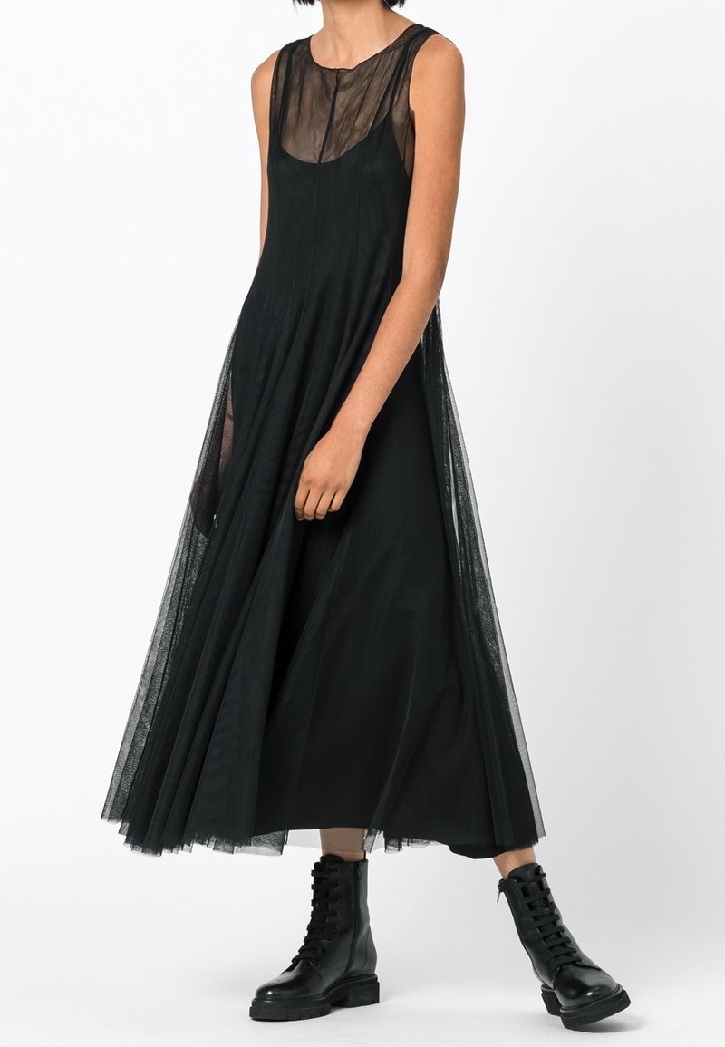 Personne portant une robe midi noire sans manches, transparente et superposée, avec un corsage ajusté et des bottines noires à lacets sur fond blanc.