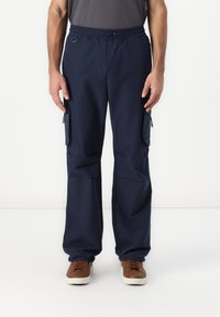Navy cargo pants with an elastic waistband, two side pockets with zippers, loose fit, and a smooth, lightweight fabric. Brown shoes visible.