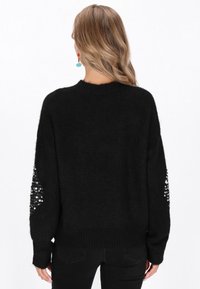 Schwarzer Pullover mit lockerer Passform, weichem Material, Rundhalsausschnitt und Paillettenakzenten an den Ärmeln. Getragen mit schwarzen Hosen.