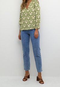 Femme portant un blouse à imprimé géométrique vert et crème, un jean droit bleu clair et des sandales à talons carrés marron, se tenant contre un mur blanc.