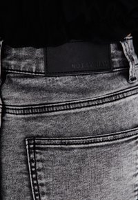 Gros plan sur la poche arrière d'un jean denim gris et une étiquette de marque noire cousue au-dessus avec "NOISY MAY" en relief.