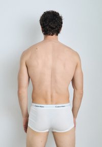 Bijele pamučne bokserice s crnim pojasom na kojem je logo "Calvin Klein", nosi osoba viđena s leđa na svijetloj pozadini.