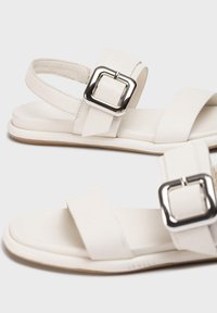 Sandales en cuir blanc avec deux larges lanières, une boucle carrée en argent et une semelle plate avec une finition texturée subtile.