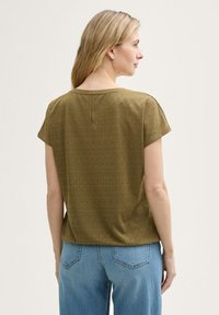 Top olive, cu mânecă scurtă, cu model din ochiuri, decolteu rotund și detaliu cu nasture la spate, asortat cu blugi din denim albastru deschis.