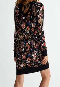 Květinové šaty s dlouhým rukávem v černé tkanině, s barevnými květinovými vzory, výstřihem do V a detailem mašle na zadní straně. Žebrované manžety a lem.