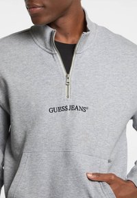 Pelēks ar rāvējslēdzēju aizdaramais džemperis, izgatavots no mīksta auduma, ar augstu apkakli, priekšējo kabatu un melnu izšuvumu "GUESS JEANS" logo.