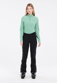 Maglione di pile verde menta a maniche lunghe con collo a zip, abbinato a pantaloni da sci neri. Tessuto resistente, design aderente, con scarponi da sci in vista.