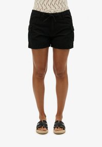 Pantalones cortos de carga negros con cintura de cordón, múltiples bolsillos laterales y un ajuste relajado, combinados con sandalias negras de suela trenzada.