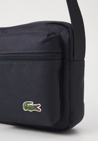 Borsa a messenger in tessuto nero con una tasca con cerniera e una superficie texturizzata. Presenta un logo di coccodrillo verde sul davanti. Tracolla regolabile.