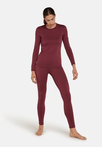 Donna che indossa un top aderente bordeaux a maniche lunghe e leggings abbinati, in piedi a piedi nudi su uno sfondo bianco, guardando in basso.