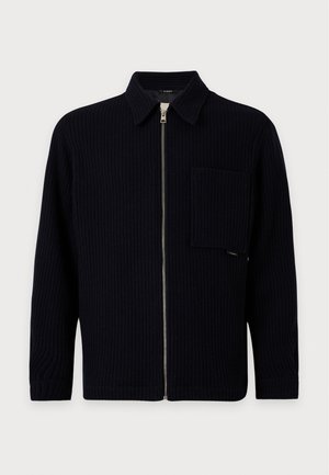 Jeune homme portant une veste noire texturée zippée, une chemise grise, un jean large foncé et des chaussures en cuir noires, debout devant un fond uni.