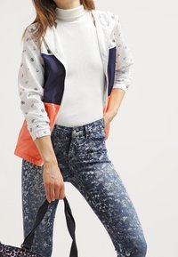 Farverig vindjakke med hvide, marineblå og koralfarvede sektioner, prydet med anker mønstre. Båret over en hvid rullekrave og parret med slidte blå jeans.