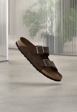 Sandalo in camoscio marrone con due fibbie metalliche regolabili e soletta in sughero, mostrato fluttuante in ambienti interni su pavimento liscio con sfondo sfocato.