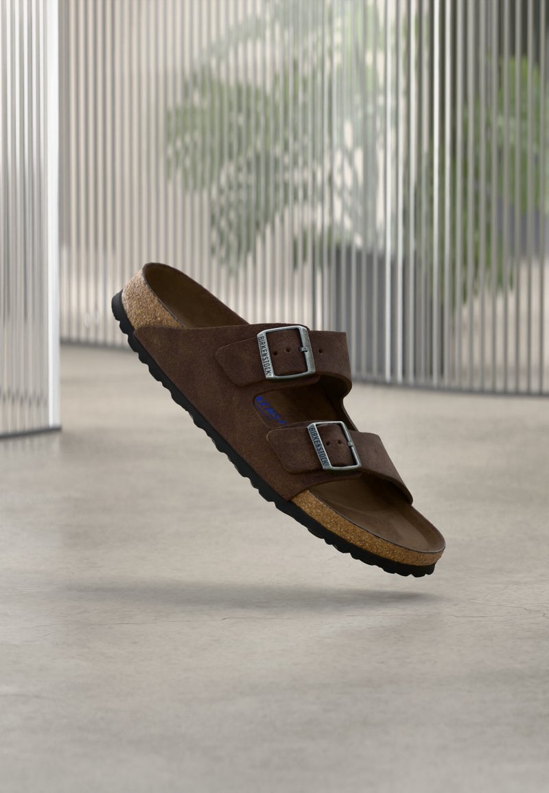 Sandale en daim marron avec deux boucles métalliques réglables et une semelle en liège, présentée flottante à l’intérieur sur un sol lisse avec un arrière-plan flou.
