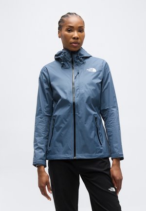 Femme portant une veste imperméable bleue avec capuche et un pantalon noir, tous deux de la marque "The North Face", debout devant un fond blanc uni.