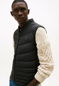 Chaleco acolchado negro con costuras verticales, cierre de cremallera y un suéter de punto beige con patrón texturizado. Cuello simple y sin marcas visibles.