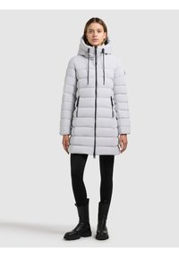 Ljusgrå vadderad jacka med horisontella sömmar, dragkedja fram, dragsko i huvan och två sidofickor. Bärs över svarta leggings och stövlar.