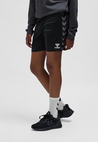 Zwarte sportshorts met een gestructureerde tailleband, met een chevronpatroon langs de zijkanten, gecombineerd met zwarte sportschoenen en witte sokken.