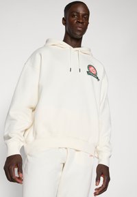 Krémsová mikina z jemného materiálu, s okrúhlym logom a šnúrkou, v kombinácii s rovnako krémsovými nohavicami.