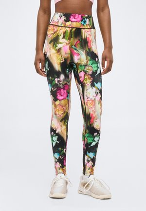 Leggings mit lebhaftem mehrfarbigem Blumenmuster, getragen von einer stehenden Person, kombiniert mit weißen Sportschuhen.