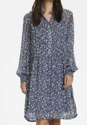 Frau trägt ein blaues, geblümtes Kleid mit langen Ärmeln, gesammelten Bündchen und vorderen Biesen, steht vor einem weißen Hintergrund.