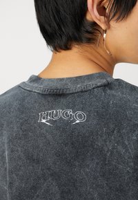 Camiseta de algodón negra y desgastada con cuello redondo; presenta el logo bordado en blanco "HUGO" en la parte de atrás, mostrando un aspecto texturizado y viejo.
