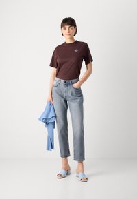 Braunes Baumwoll-T-Shirt mit kleinem blauen Grafikdesign, hellblauer Strickpullover über dem Arm, hellblaue gerade Jeans in heller Waschung und blassblaue Slidesandalen.