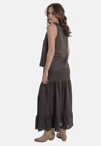 Bruine linnen mouwloze top en gelaagde maxi rok met een gerimpelde zoom. Model draagt beige enkellaarsjes en staat tegen een effen achtergrond.