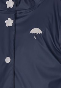 Námornícky plášť do dažďa s hviezdicovými gombíkmi a strieborným logom dáždnika na hrudi, vyrobený z hladkého, vodoodolného materiálu.