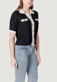 Maglione nero a maniche corte con colletto bianco, tessuto testurizzato, chiusura con bottoni e due tasche sul petto. Indossato con jeans aderenti blu chiaro.