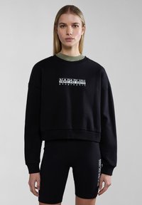 Sudadera negra corta con puños acanalados, que cuenta con un diseño de logo blanco. Se lleva sobre una camiseta verde oscura y se combina con pantalones cortos negros.