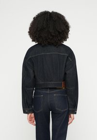 Ciemnoniebieska krótka kurtka jeansowa z dopasowaną talią, szerokimi rękawami i skórzaną metką z logo. Tekstura jest gładka z widocznymi detalami przeszyć.