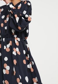Granatowa sukienka z zaznaczoną talią, ozdobiona dużymi kropkami w kolorze brzoskwiniowym, białym i czarnym. Długie rękawy z marszczonymi mankietami.