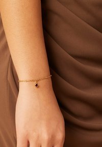 Gouden kettingarmband met een kleine rode edelsteenhanger, gedragen om een pols tegen een bruine stofachtergrond. Subtiele textuur op de huid.