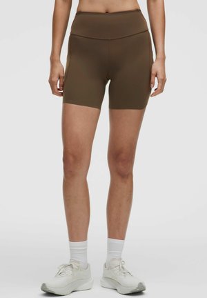Kvinde iført brune højtaljede tætsiddende shorts, hvide ankelsokker og hvide sportssko, stående mod en ensfarvet baggrund.