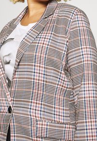 Kobieta nosząca wielokolorową marynarkę w kratę na białym T-shircie z czarno-białą grafiką twarzy.