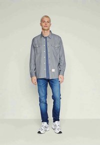 Gråstripet skjorte med knapp, to brystlommer, brukt over en marineblå t-skjorte, kombinert med blå jeans og hvite joggesko.