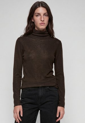 Giovane con capelli castani lunghi che indossa un dolcevita marrone scuro e jeans neri, in piedi davanti a uno sfondo grigio chiaro uniforme.