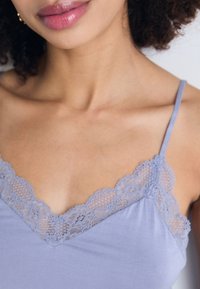 Camisole lilas clair avec une garniture en dentelle le long du décolleté en V, des bretelles fines et une texture de tissu lisse. La dentelle détaillée ajoute une touche décorative.