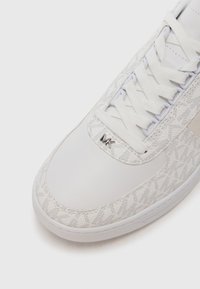 White sneaker with a textured fabric upper featuring a subtle pattern, round toe, and flat white laces; includes a silver logo accent.