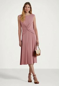 Donna in un abito midi senza maniche di un rosa polveroso con dettaglio a nodo frontale, che tiene una borsa piccola bicolore e indossa sandali con tacco alti marroni.