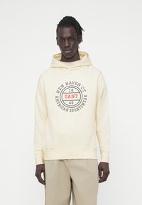 Φούτερ με κουκούλα σε εκρού χρώμα, κατασκευασμένο από απαλό ύφασμα, με κυκλικό γραφικό σε μαύρο και κόκκινο που αναγράφει "NEW HAVEN CT," "GANT" και "AMERICAN SPORTSWEAR."